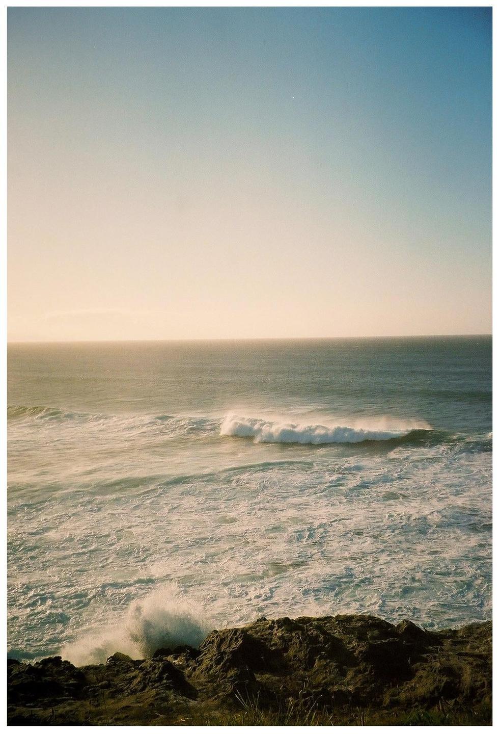 A large wave breaks on the shore of a rocky coastline under a clear sky.
