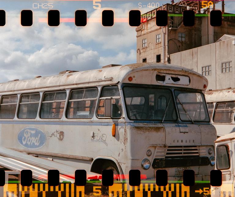 A weathered white bus with a Ford logo on its side is parked in front of a building under a cloudy sky.