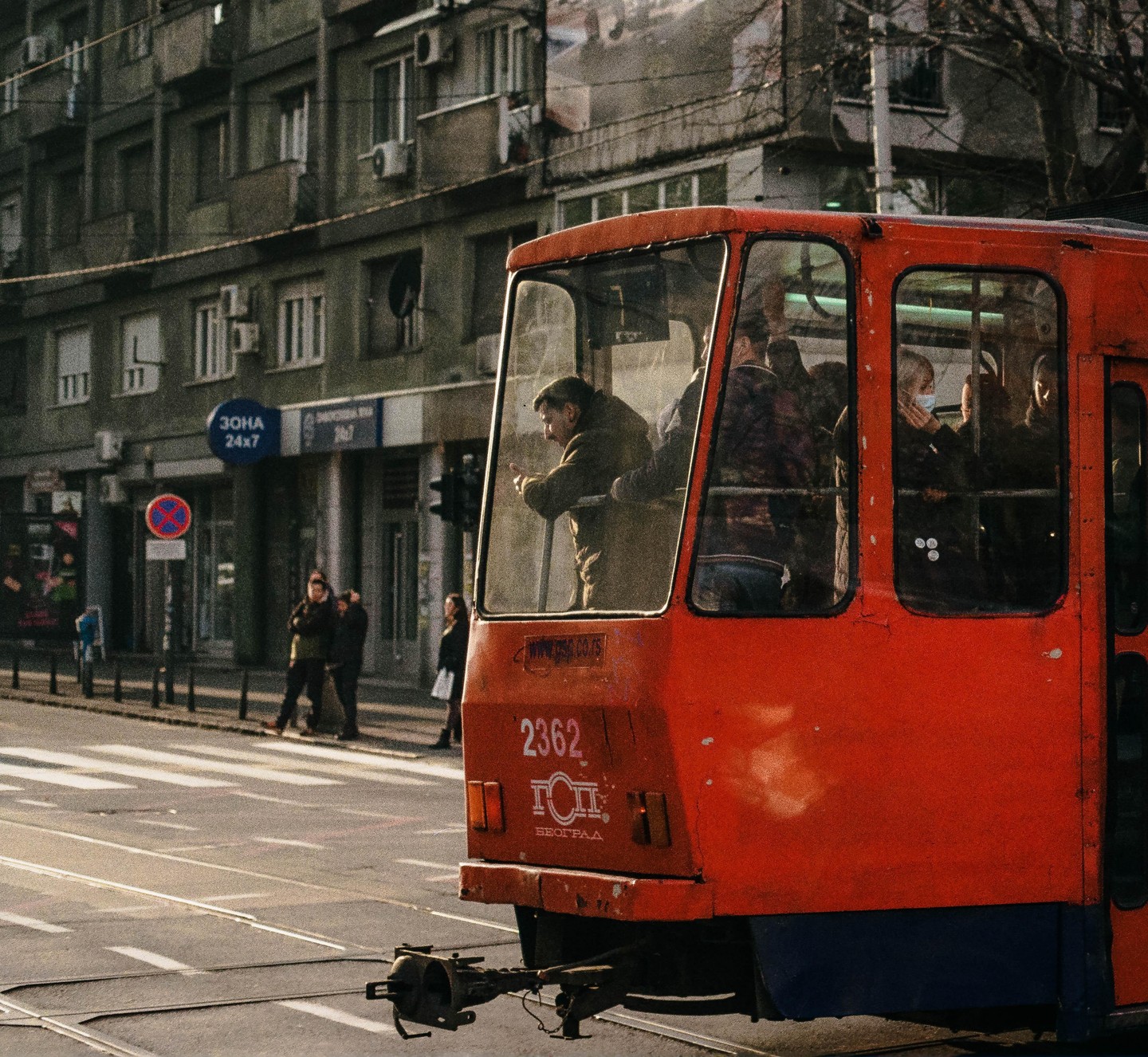 A red tram with passengers inside is stopped on a city street with buildings in the background.