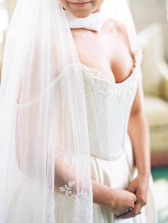 A woman in a white wedding dress and veil stands with her hands clasped.