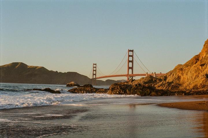 The Golden Gate Bridge is visible in the distance from a rocky beach with waves crashing on the shore.