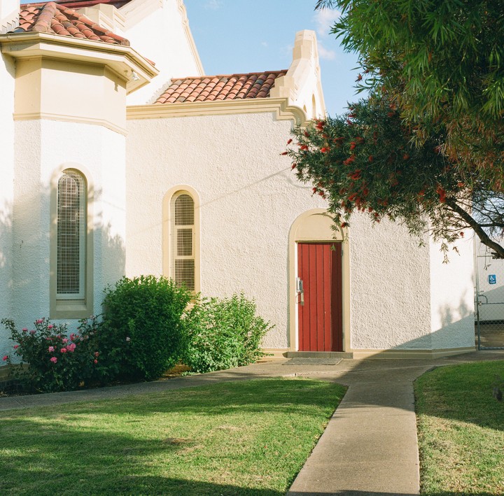 A red door is visible on a white building with a tiled roof, next to a green lawn and bushes.