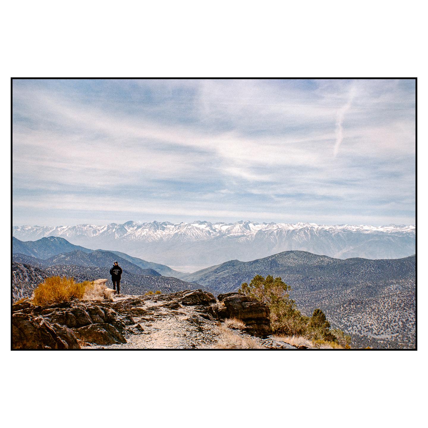 A person stands on a rocky outcrop overlooking a vast mountain range with snow-capped peaks under a cloudy sky.
