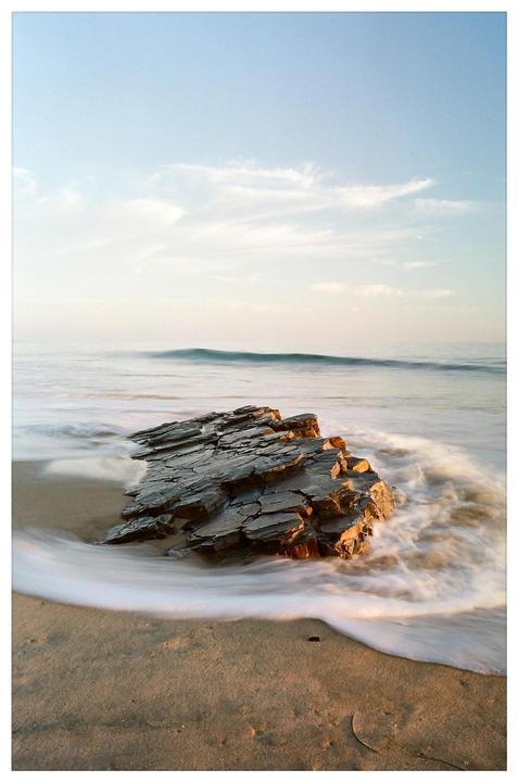 A large rock formation sits on a sandy beach as waves crash around it under a clear sky.