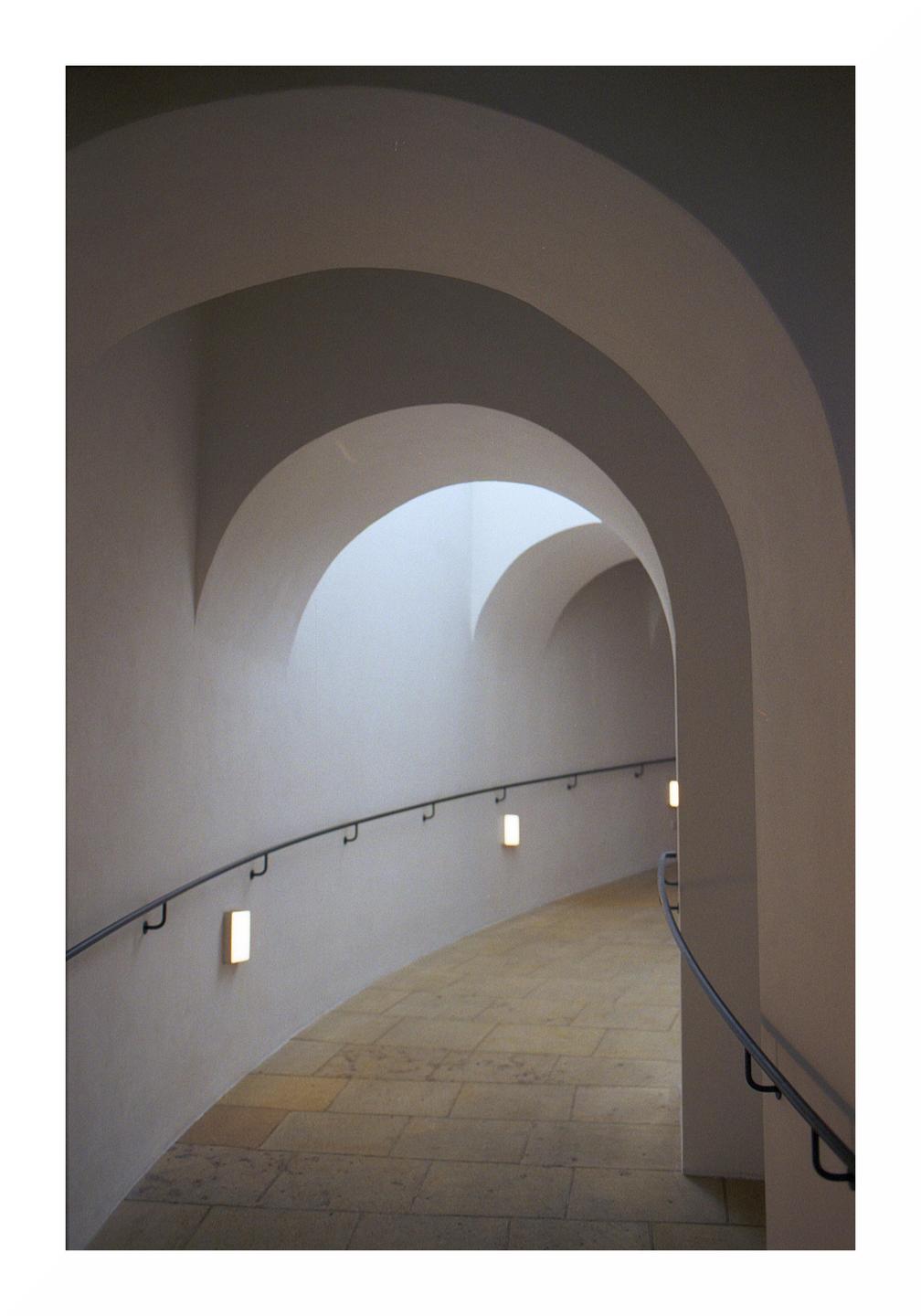 A curved hallway with a handrail and wall lights leads to an arched opening with light coming through.