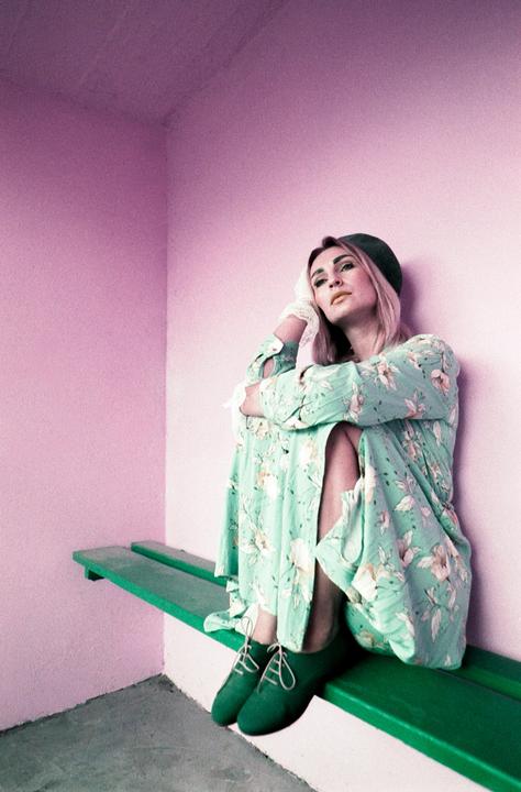 A woman wearing a floral dress, green shoes, and a green beret sits on a green bench against a pink wall.