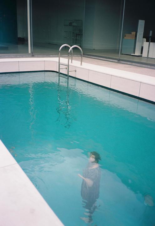 A woman stands in a swimming pool with a ladder and a building with large windows in the background.