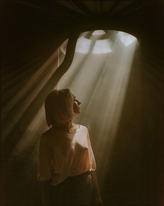 A woman stands in a dark room with rays of light shining down from an overhead window.