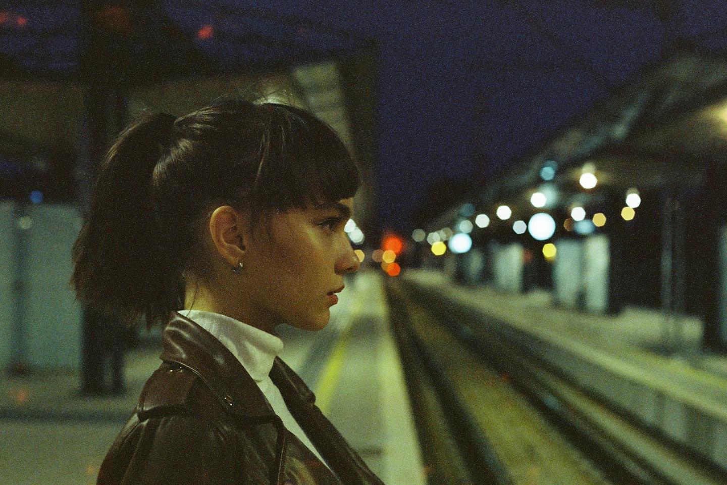 A woman with a ponytail and bangs stands on a train platform at night, looking to the right.
