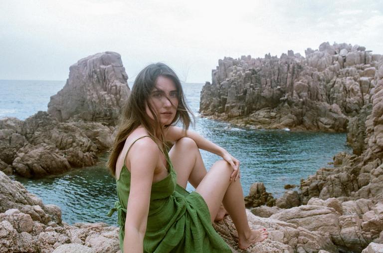 A woman in a green dress sits on a rocky shore overlooking the sea.