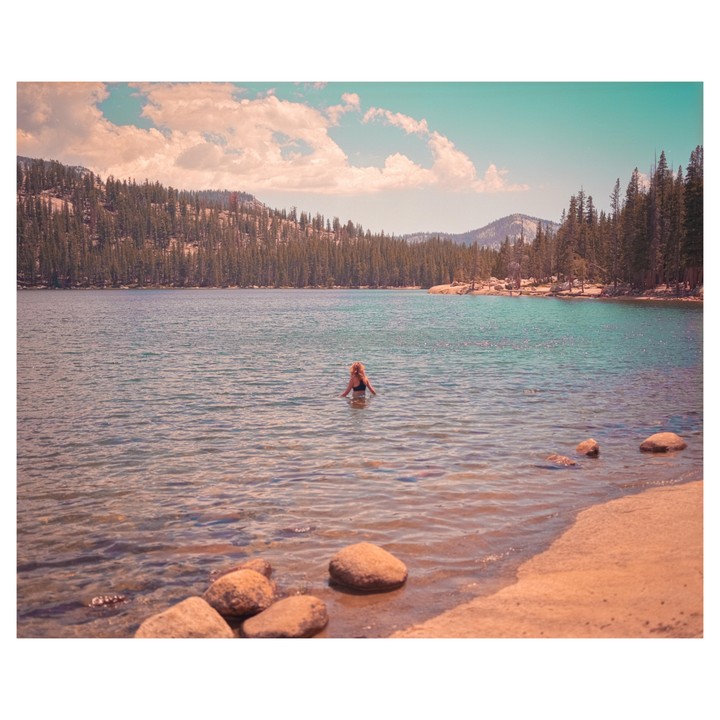 A woman stands in a lake surrounded by a forest and mountains under a cloudy sky.