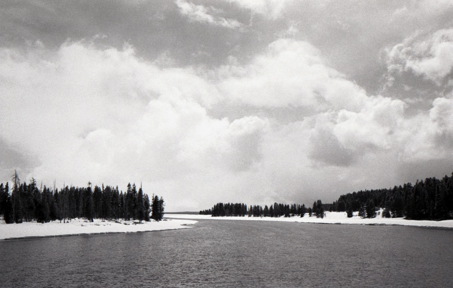 A wide river flows between snow-covered banks lined with pine trees under a cloudy sky.