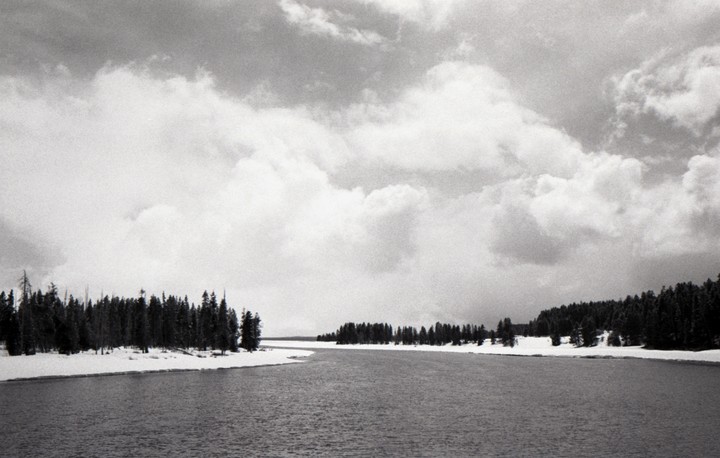 A wide river flows between snow-covered banks lined with pine trees under a cloudy sky.