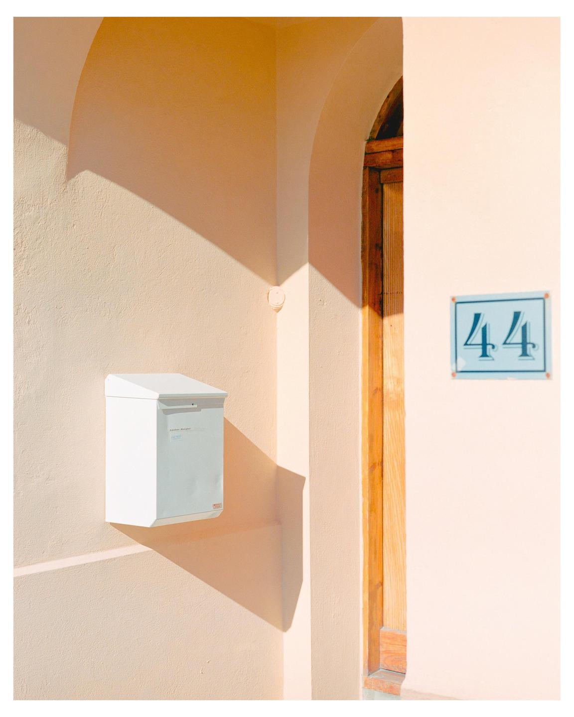 A white mailbox is mounted on a peach-colored wall next to a wooden door frame and a blue house number sign.