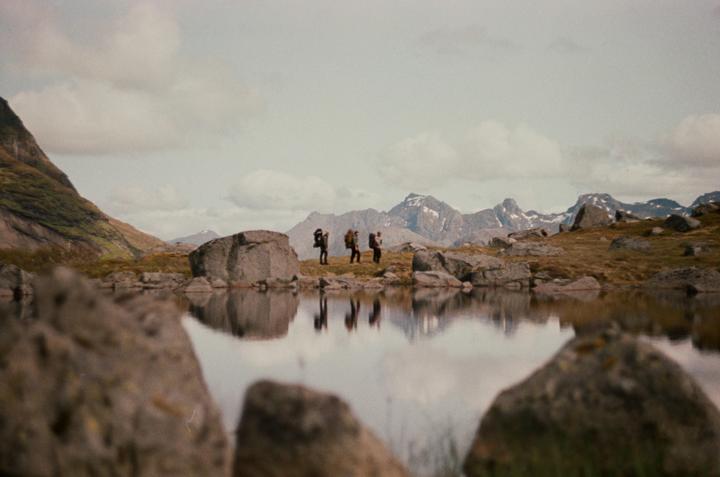 Three people with backpacks stand by a lake with mountains in the background.