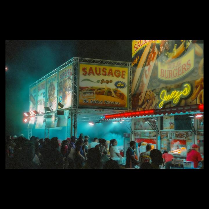 A crowd of people gathers around a brightly lit food stall selling sausages and burgers at night.