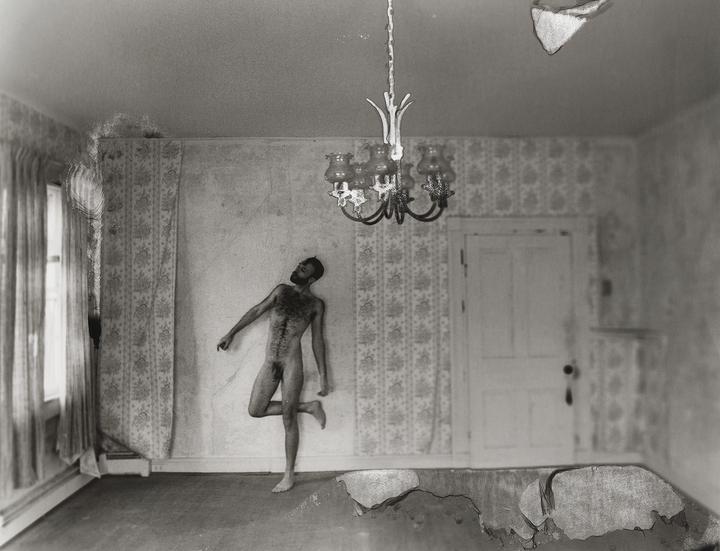 A nude man stands in a room with patterned wallpaper, a window, and a chandelier.