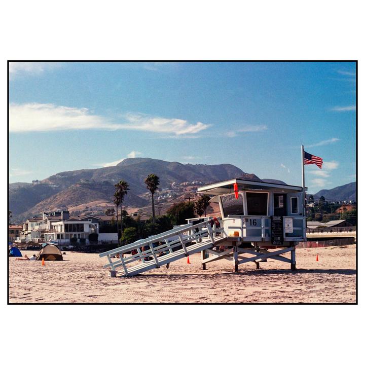 A lifeguard tower with an American flag stands on a sandy beach with mountains and houses in the background.