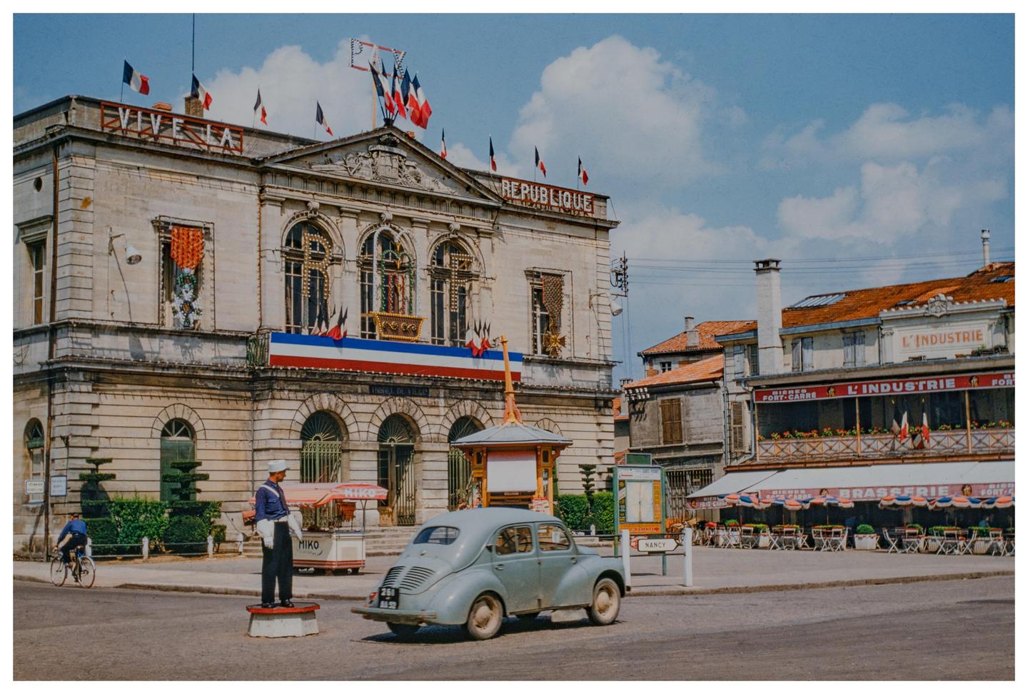 A street scene in France shows a building with "REPUBLIQUE" written on it, a vintage car, and a man on a bicycle.