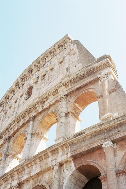 The Colosseum in Rome is captured from a low angle, showing its weathered stone and brick arches against a clear sky.