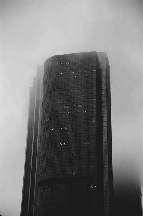 A tall, curved skyscraper with many windows rises into a hazy sky, with other buildings faintly visible behind it.