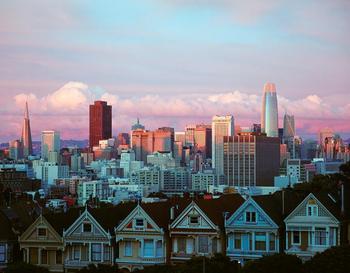 A row of Victorian houses sits in the foreground with a city skyline and pink clouds in the background.
