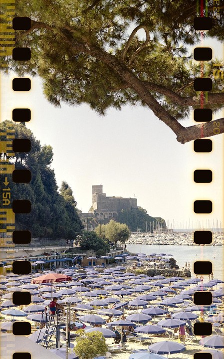 A beach with many blue umbrellas and people is visible, with a castle and a harbor full of boats in the background.