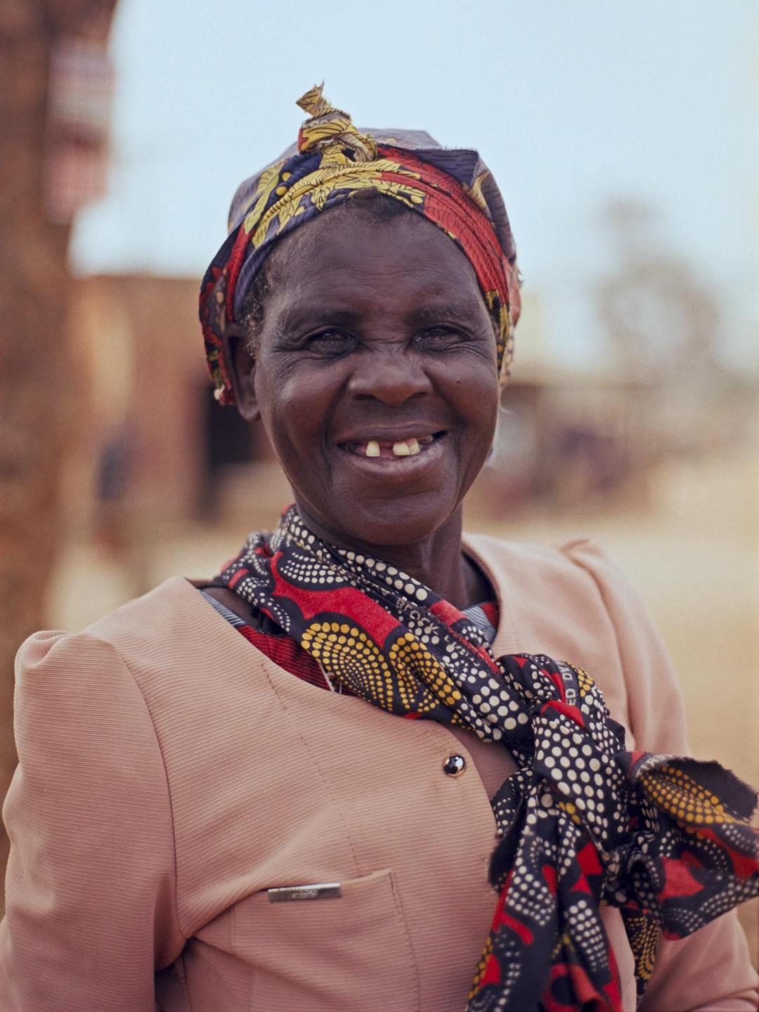 A woman with a colorful headscarf and scarf smiles at the camera.