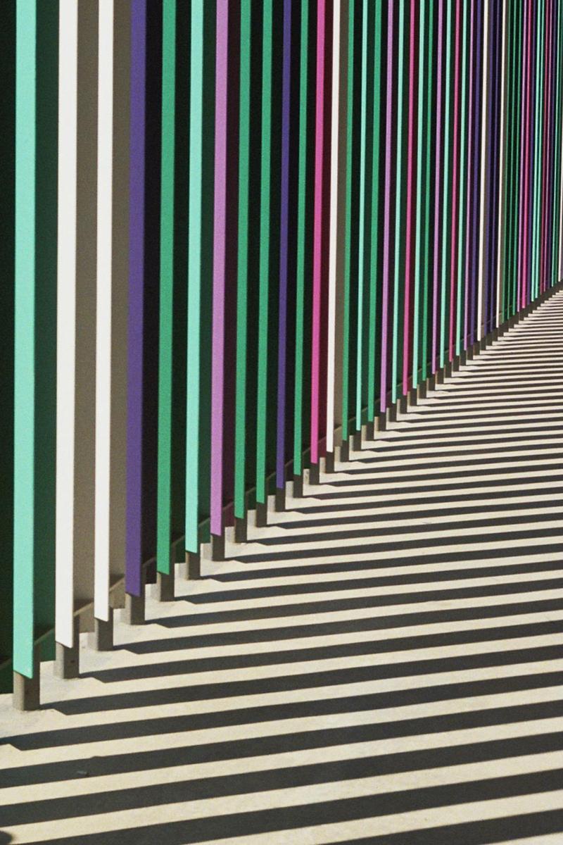 A colorful striped wall casts long, parallel shadows onto the ground.