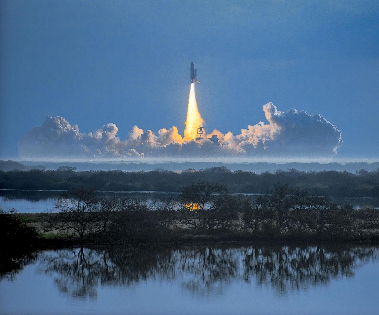 A space shuttle launches into a blue sky, leaving a trail of fire and smoke above a body of water and trees.