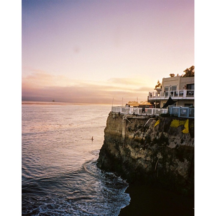 A person swims in the ocean next to a cliff with a house on top at sunset.