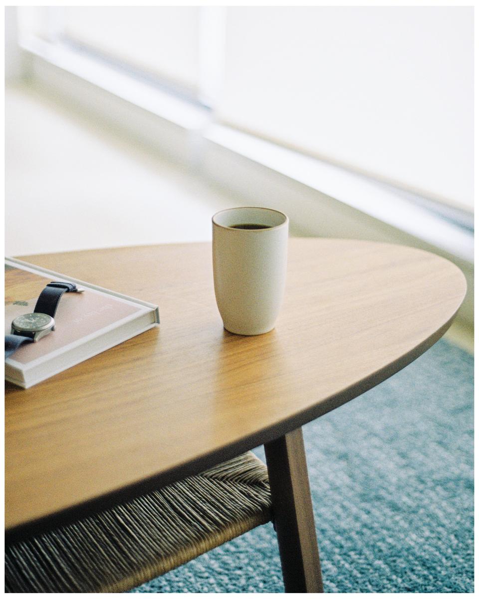 A cup of coffee sits on a wooden table next to a book and a watch.