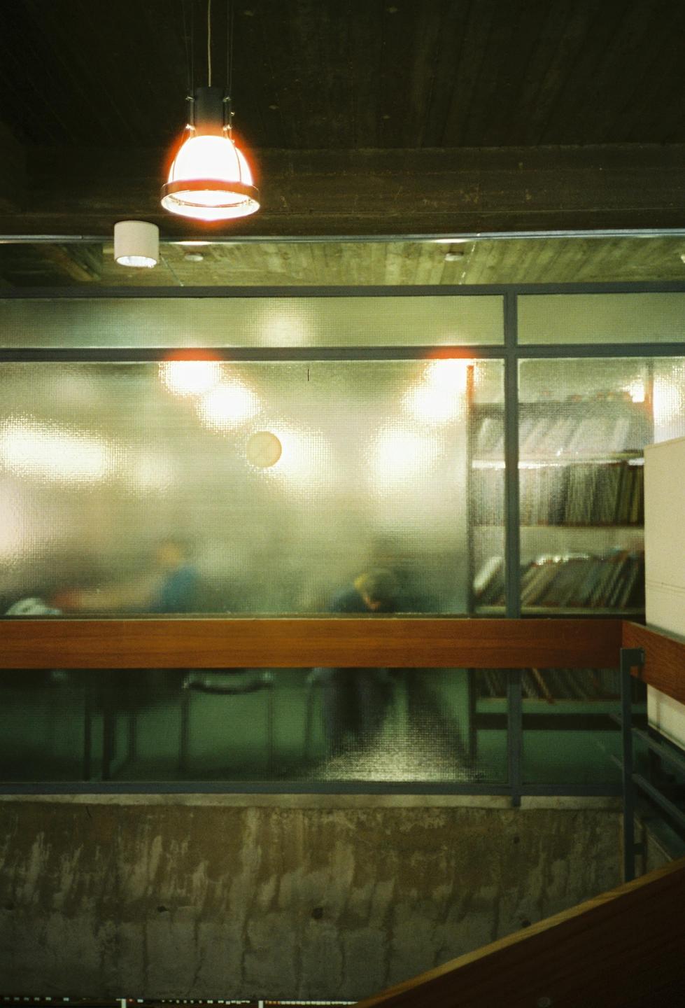 A hanging lamp illuminates a textured glass wall, behind which blurred figures and shelves of records are visible.