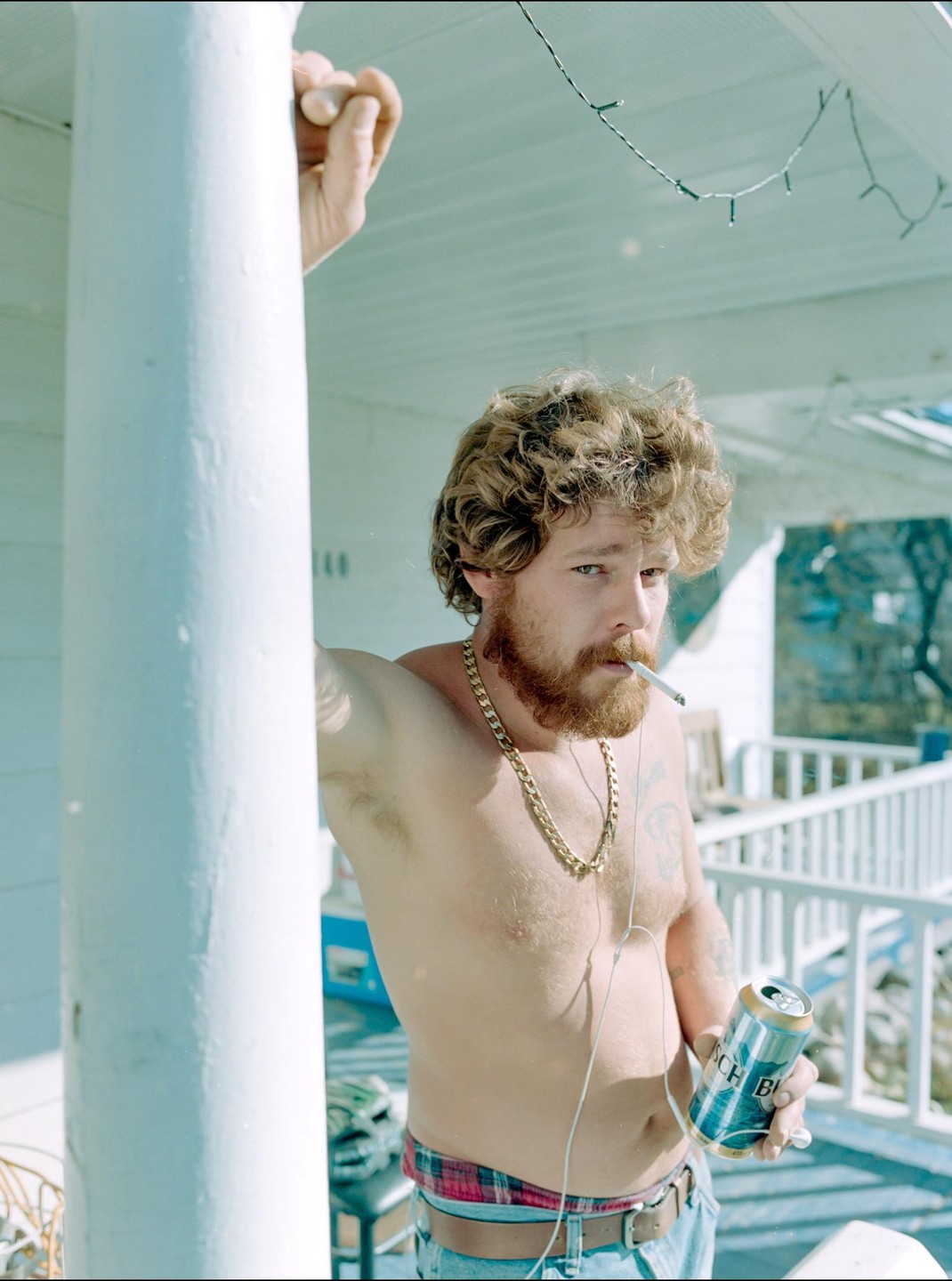 A shirtless man with a beard and curly hair holds a beer can and a cigarette while leaning against a white pillar on a porch.