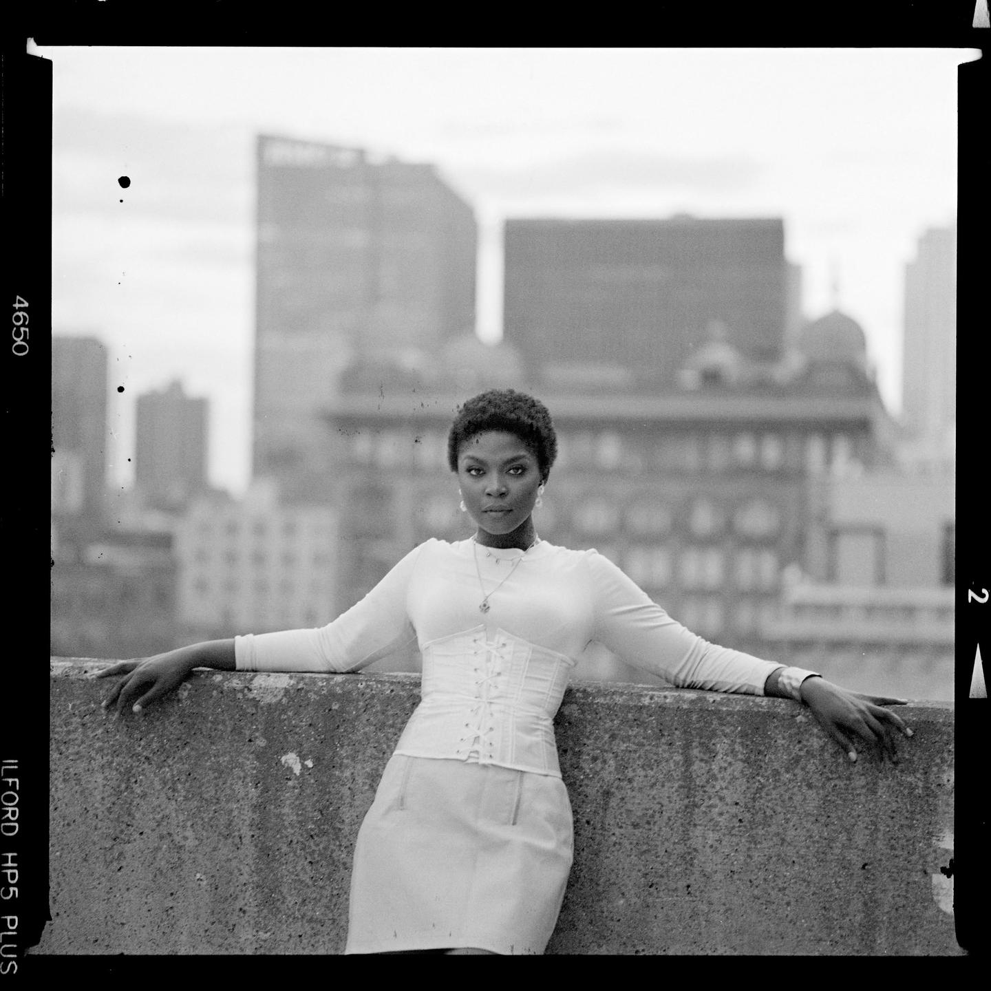 A woman in a white top and skirt leans against a concrete wall with a city skyline in the background.