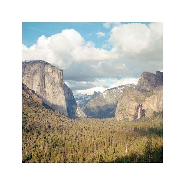 A wide valley filled with pine trees is surrounded by tall mountains, with a waterfall visible on the right side and a cloudy sky above.