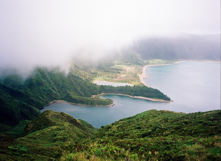 A misty landscape shows a lake surrounded by green hills and a narrow strip of land.