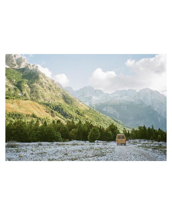 A yellow van drives on a gravel road through a valley with mountains and trees.