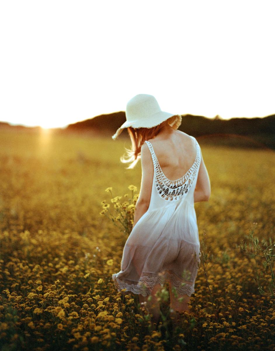 A woman in a white dress and hat walks through a field of yellow flowers at sunset.