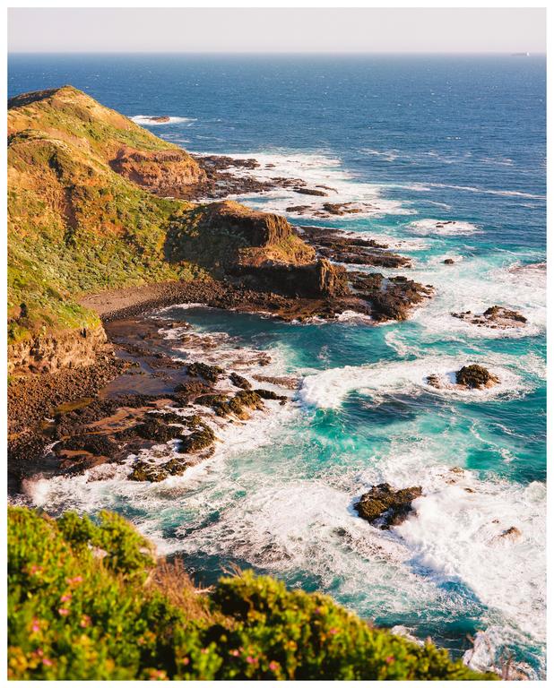 A high-angle landscape view shows a rocky coastline with green hills meeting a turquoise ocean with white waves.