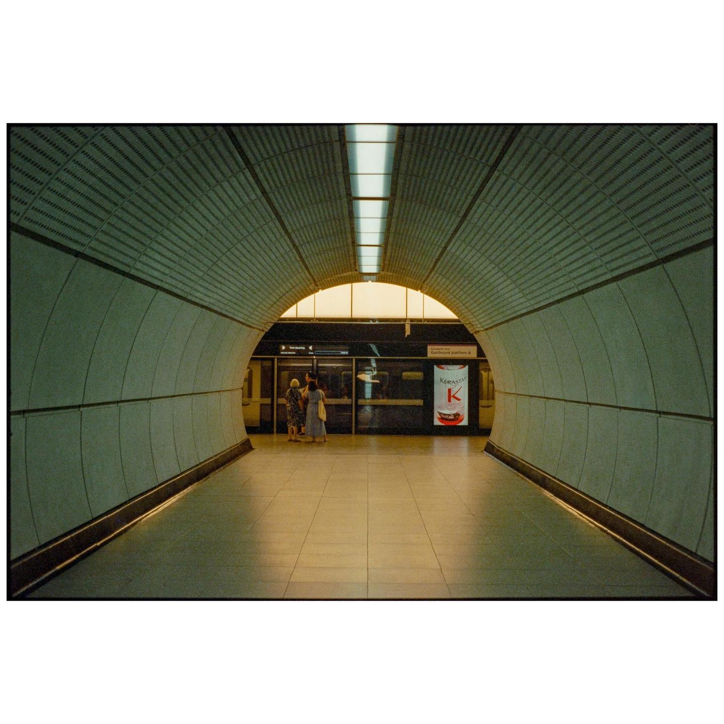 Two women stand on a subway platform in a long, arched tunnel.