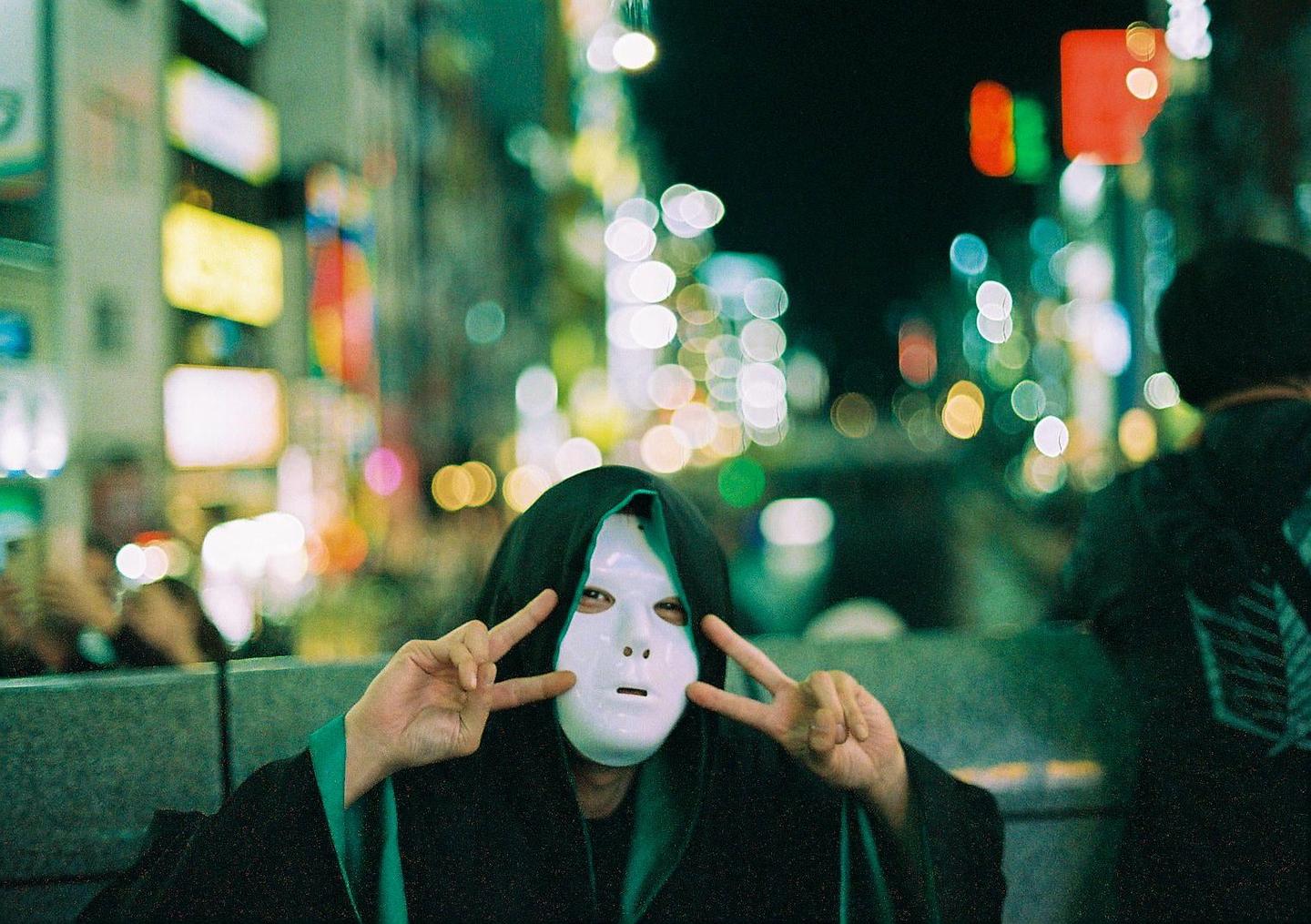 A person wearing a white mask and a dark hooded robe makes a peace sign with both hands at night on a city street.