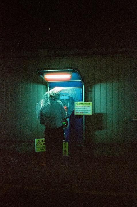 A person holding an umbrella stands in front of an ATM at night in Hiroshima.