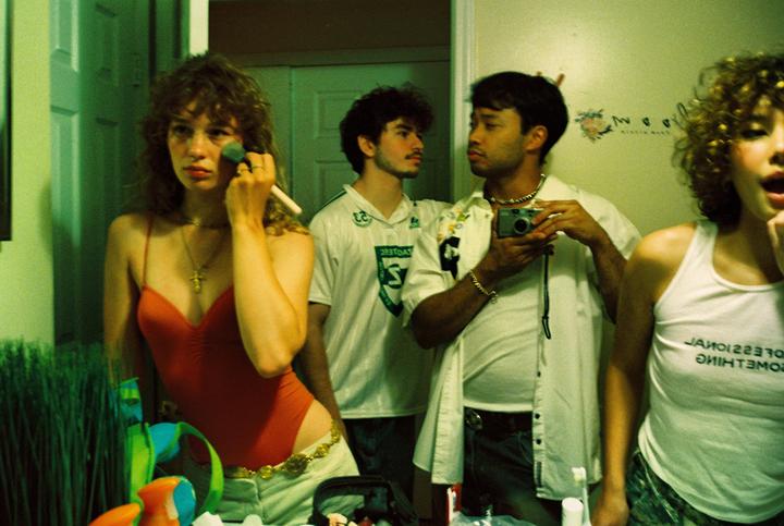 A woman applies makeup while three men are reflected in a mirror, one holding a camera.
