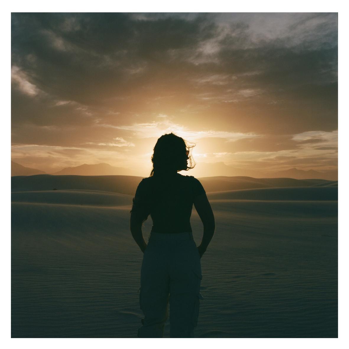 A woman stands silhouetted against a sunset over sand dunes and mountains.
