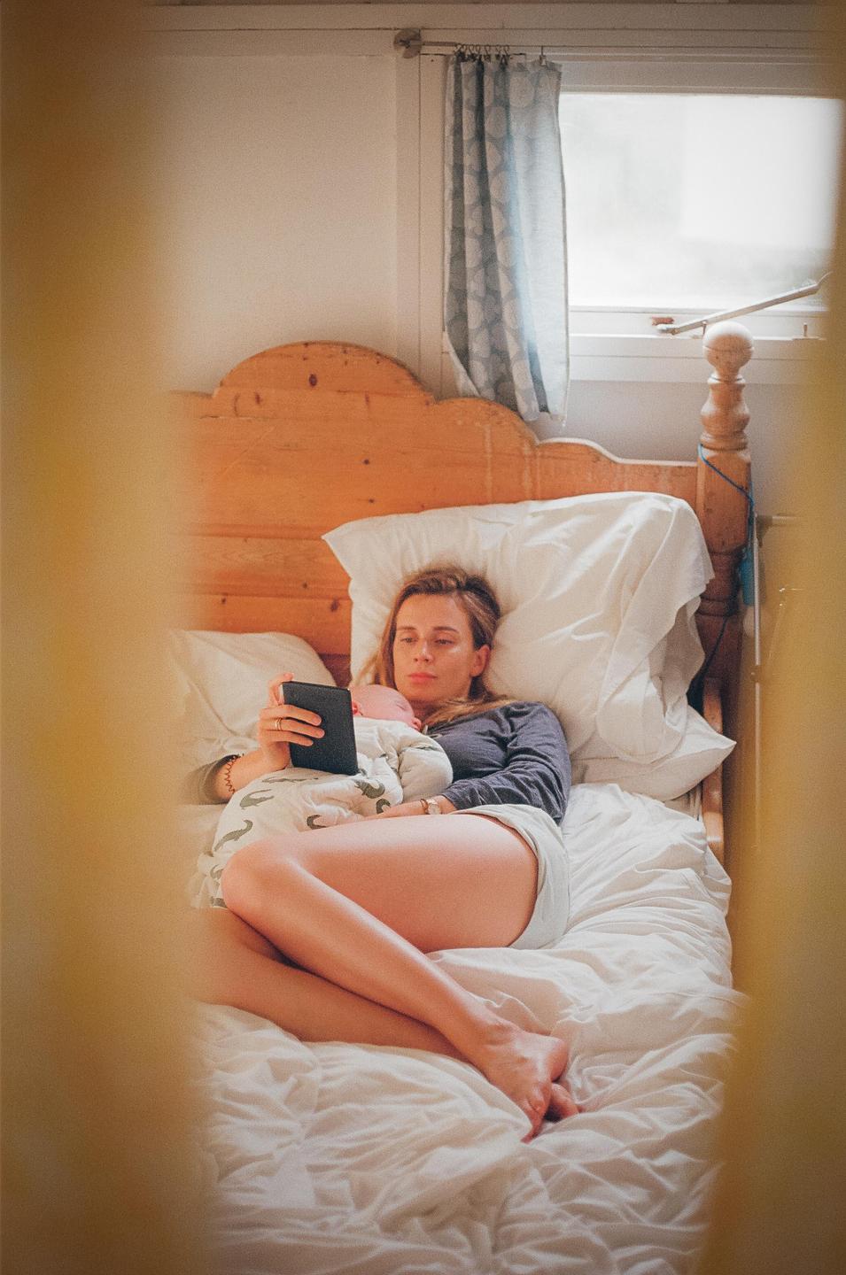 A woman lies in bed with a baby on her chest while looking at an e-reader.