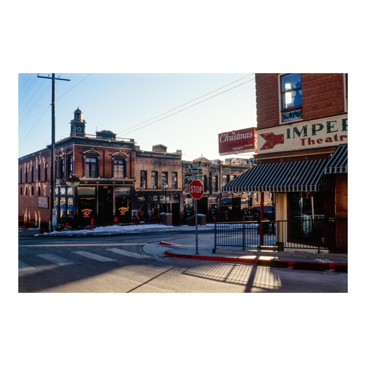 A street corner in a historic town features brick buildings, a stop sign, and a crosswalk with long shadows.