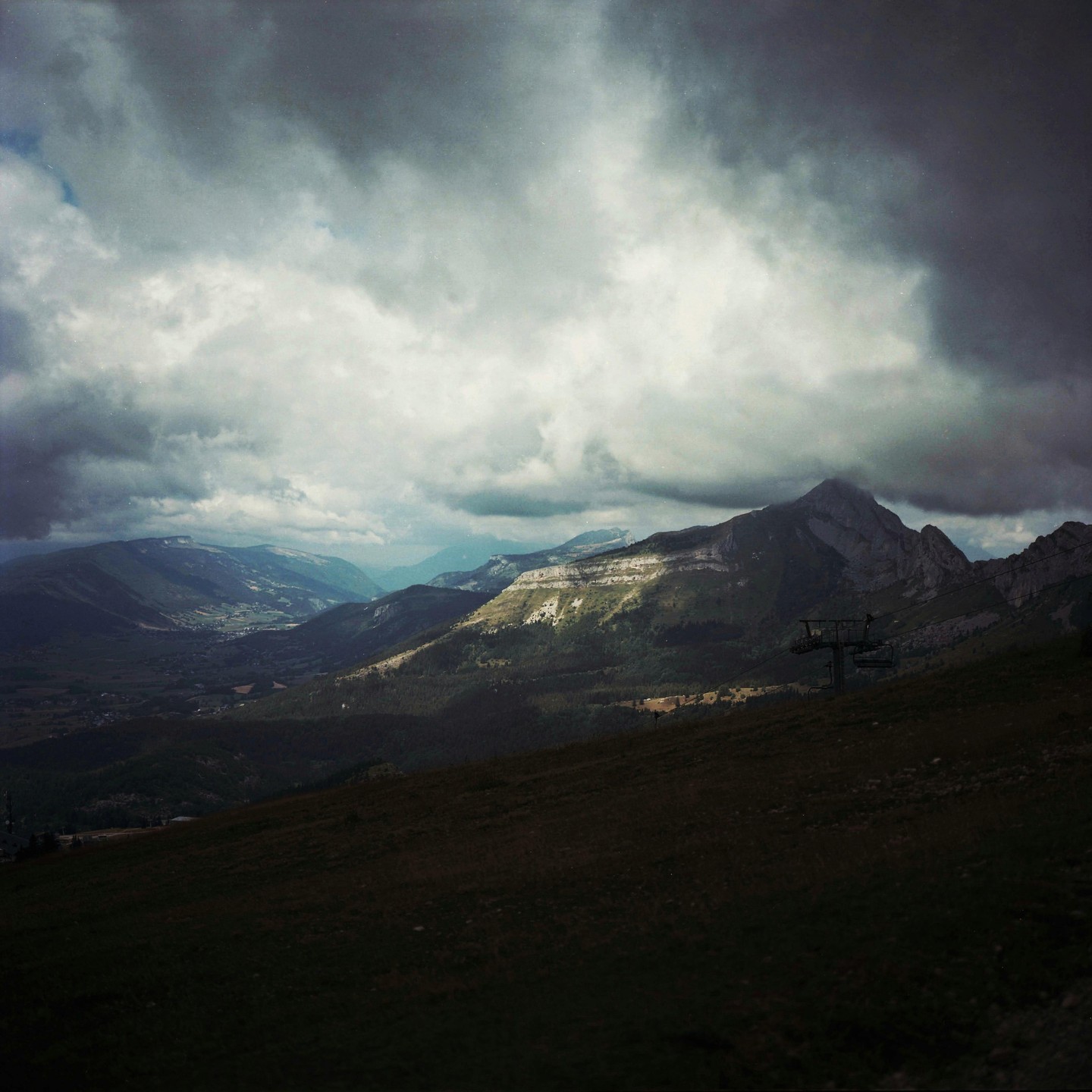 A dramatic mountain landscape shows a valley and distant mountains under a cloudy sky, with a ski lift in the foreground.
