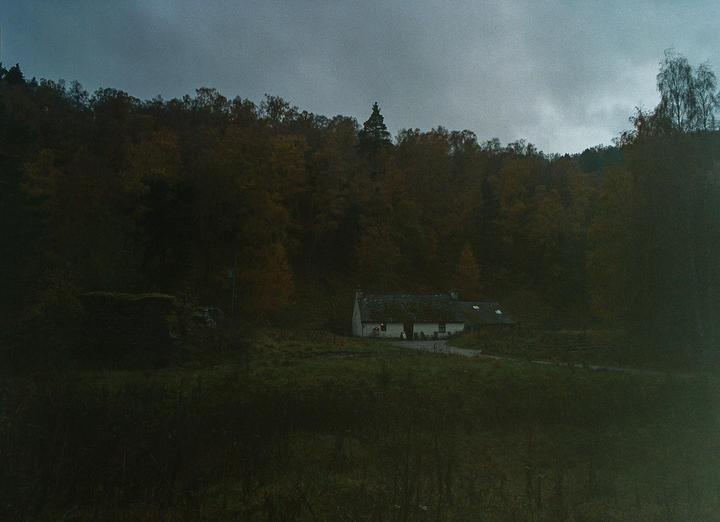A small white house with a dark roof sits in a grassy field at the base of a forest under a cloudy sky.