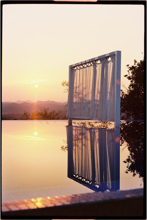 A sunset over mountains is reflected in a swimming pool next to a curtained structure.